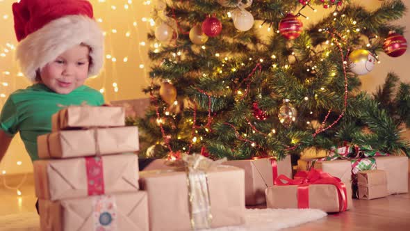 Little Child Kid Boy Green Shirt and Red Santa Hat Under Fir Tree with Presents alt