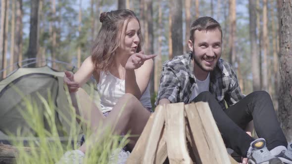 Portrait Handsome Young Man Kindling a Fire in the Forest While Beautiful Young Woman Sitting Near alt