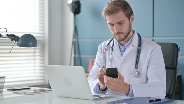 Doctor Using Smartphone While Working on Laptop in Clinic alt
