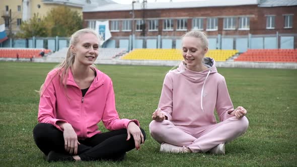 Two Young Fit Smiling Twins in Pink Sportswear Sit at Grass Field in Lotus Pose