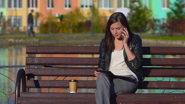 An Attractive Girl on a Bench Calls on a Smartphone and Prints on a Tablet. alt