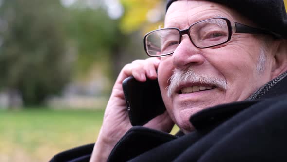 A French Senior is Talking on a Smartphone He is Sitting in a Park on a Bench alt