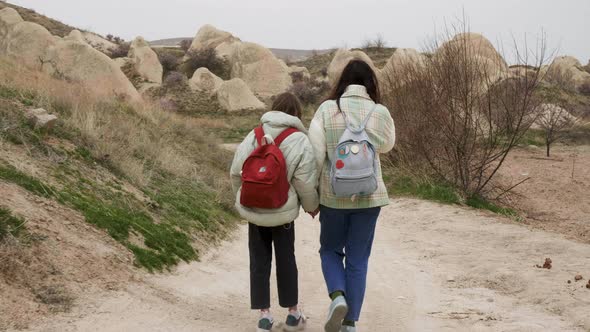 Back View of Two Unrecognizable Girls with Backpacks Walking Through Wild Road Holding Hands Against
