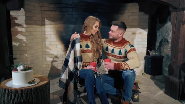 Beautiful couple is sitting near fireplace drinking tea. Man tenderly hugs woman. Intimate atmospher alt