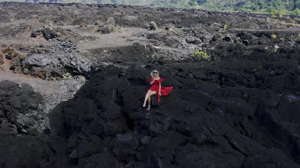 Girl in Red Dress Sitting on Solidified Black Lava Field on Volcano Batur in Bali, Indonesia. Aerial alt