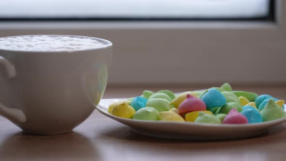 A Large Mug of Coffee with Milk or Mochacino and Colorful Sweets in a Plate on the Windowsill in