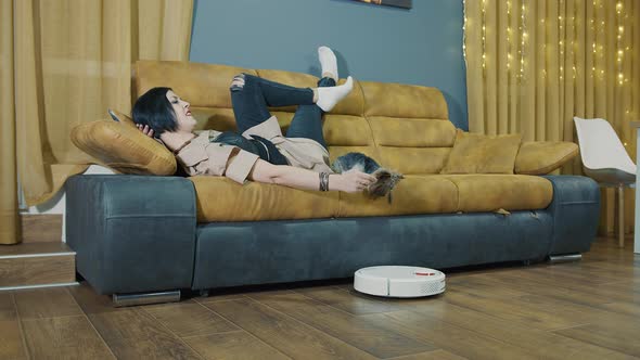 Woman Lying on the Couch with a Dog Turns on the Hand of a Robot Vacuum Cleaner alt