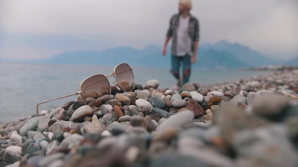 Blonde Man in a Plaid Shirt Walks to His Shades on the Pebbles alt