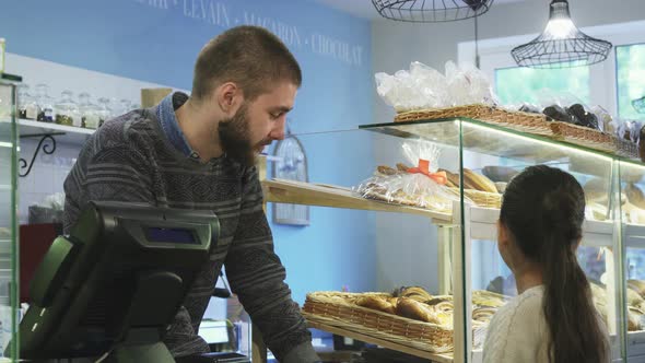 Young Male Baker Working at His Store Helping Little Girl Choosing Desserts alt