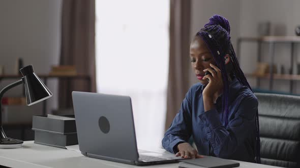 Black Woman is Calling By Mobile Phone Sitting at Working Table in Her Home Office Chatting By alt