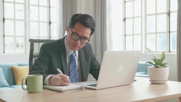 Asian Businessman Typing On Computer Then Taking Note In A Book While Working At Home alt