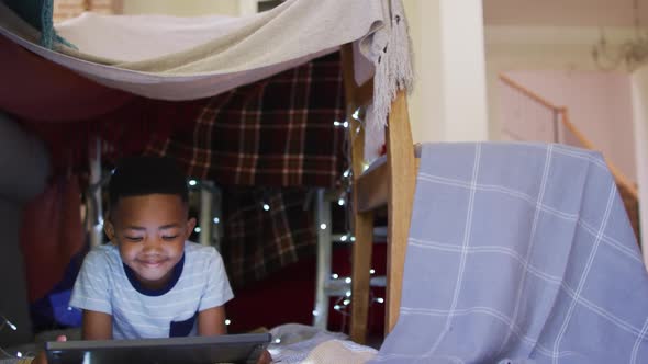 Happy african american boy lying in house made of blankets, using tablet alt