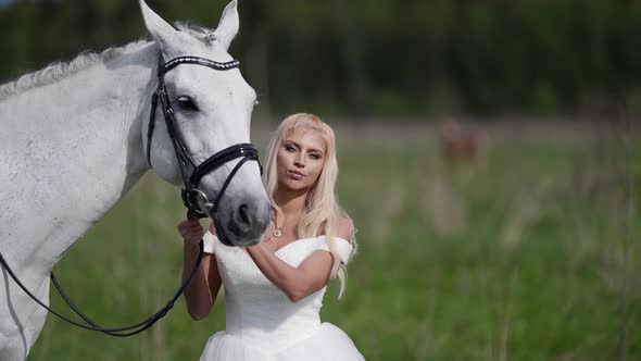 Pretty Woman in a Wedding Dress in a Pasture in the Afternoon Stroking Her Horse alt