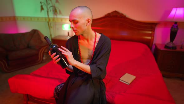 Portrait of Young Sad Feminine Man in Dress and Necklace Holding Wine Bottle Thinking alt