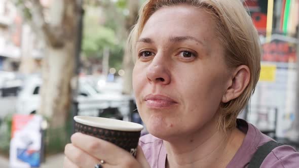 A Woman with a Thoughtful Face Holds a Mug of Hot Coffee in Her Hands in a Street Cafe in the alt
