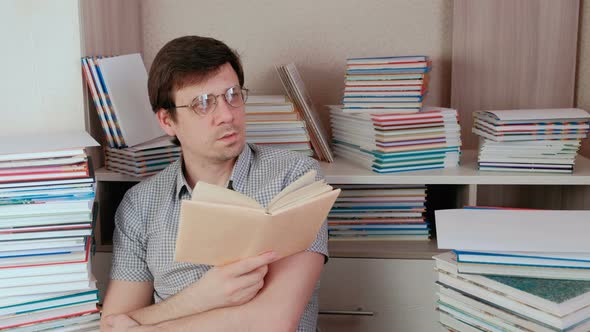 Young Brunet Man in Glasses Reads a Book and Thinks Sitting Among Books
