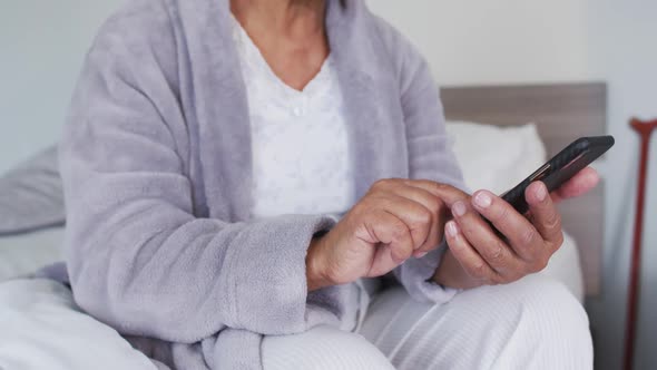 Mid section of african american senior woman using smartphone while sitting on the bed at home alt