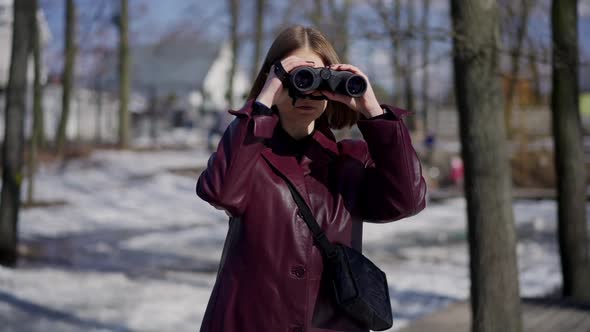 Portrait of Serious Woman with Binoculars Standing in Winter Park Looking Away alt