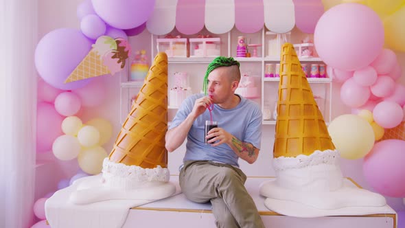 Young man with dreadlocks drinking refreshing soda, sitting on table in confectionery. alt