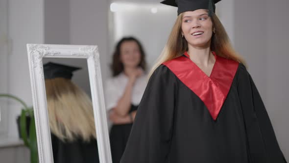 Happy Confident Young Woman in Graduation Gown and Cap Spinning Admiring Reflection in Mirror with alt