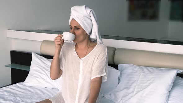 Thoughtful Girl in a Bathrobe and Towel Drinks Coffee While Sitting on the Bed
