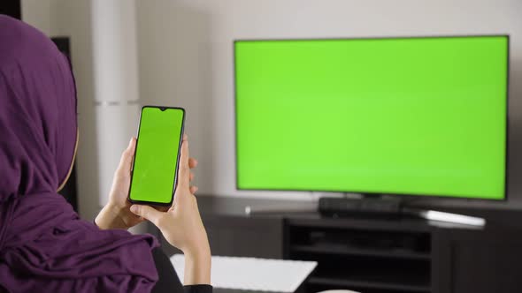 A Muslim Woman Looks at a Smartphone and TV (Green Screen) As She Sits on a Sofa in an Apartment alt