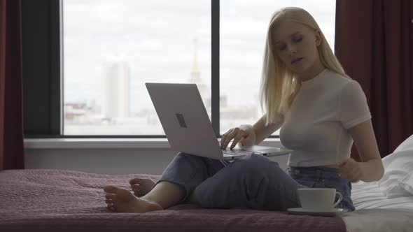 A Young Woman Freelancer Sits on a Large Bed with a Laptop and a Cup of Coffee on the Background of