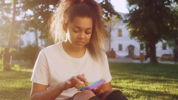 Mixed Race Teenage Girl Sitting on a Grass at the Street and Holding POP IT Fidget Toy Antistress in alt