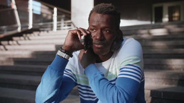 Young AfroAmerican Student Sits on the Steps and Talks on a Smartphone