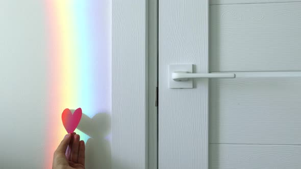 Female hand holding red paper heart on the rainbow background.