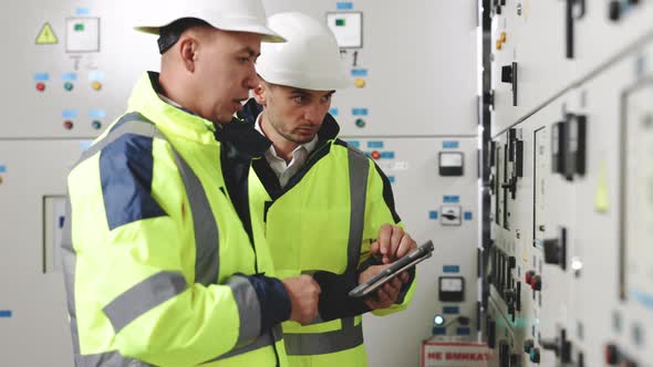 Electrical Engineers in Protective Helmet and Uniform Checking Control Panel Board with Tablet