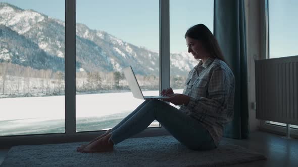 A Woman Works at Home Sitting Near the Window Using a Laptop alt