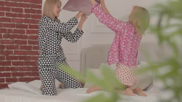 Two Laughing Blonde Caucasian Friends Fighting with Pillows at the White Bed alt