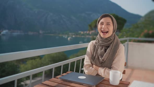 Fun Arab Young Muslim Woman in Hijab Laughing at Camera Sits Balcony Using Laptop Working Remote alt