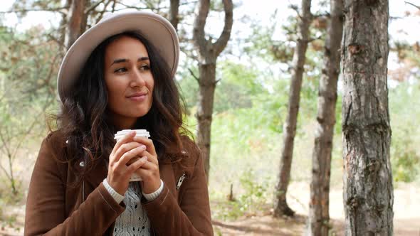 A beautiful woman on a walk among the trees drinks coffee. Food and rest. alt