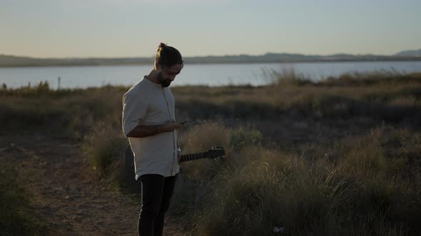 Man Guitarist Holding a Guitar and Making a Call Using a Mobile Phone While Standing on a Riverbank alt
