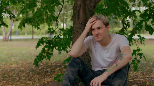 Young Man Rests and Ruffles His Hair Sitting Near Tree Trunk in Parkland alt