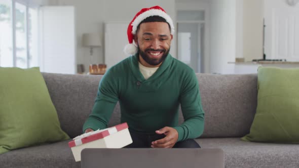 Man wearing Santa hat opening gift box while having video chat on his laptop alt