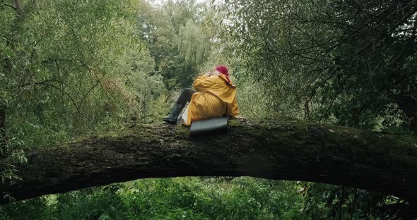 Hiker in Red Bandana and Yellow Raincoat Sits on Trunk of Fallen Tree in Forest