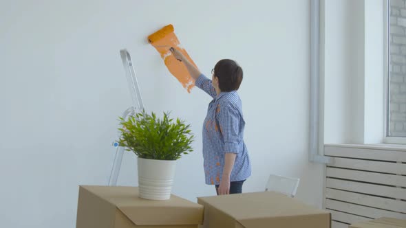 Flat Renovation Concept. Happy Middle-aged Woman Painting White Wall with Paint Roller, Orange Paint alt