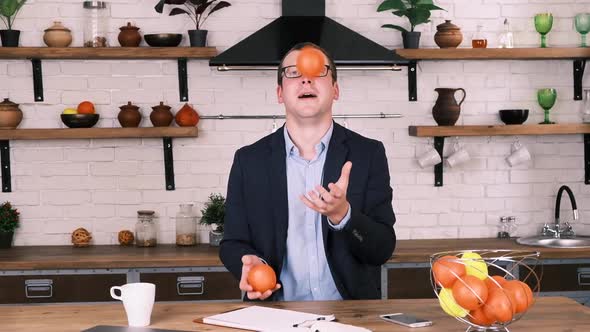 Young Cheerful Businessman in Glasses and a Business Suit Juggles with Two Oranges While Sitting in