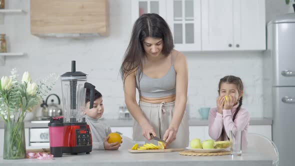 Portrait of Smiling Mother Slicing Fruits with Knife on Cutting Board in Kitchen As Children Waiting alt
