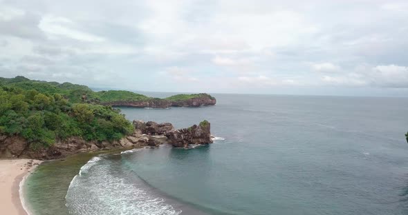 Aerial flyover coastline of Pantai during cloudy day with crashing waves against rocks on shore alt