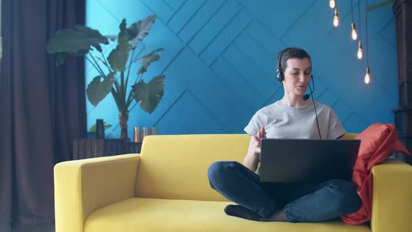 Woman with Short Hair and a Headset is Talking on Video Link a Laptop While Sitting on Yellow Sofa alt