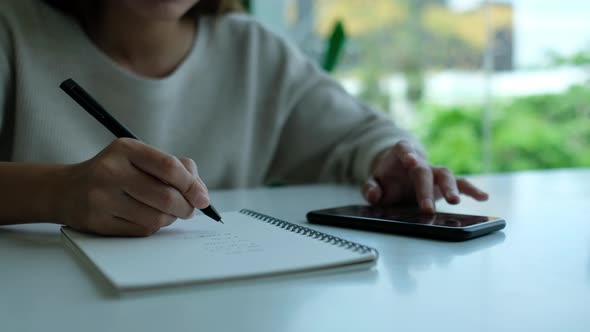 Closeup of a woman writing on a notebook while touching and scrolling on smart phone screen