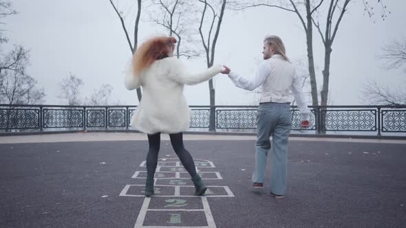 Young Caucasian Man in Elegant Clothes Holding His Girlfriend's Hand As She Jumping on the Hopscotch alt