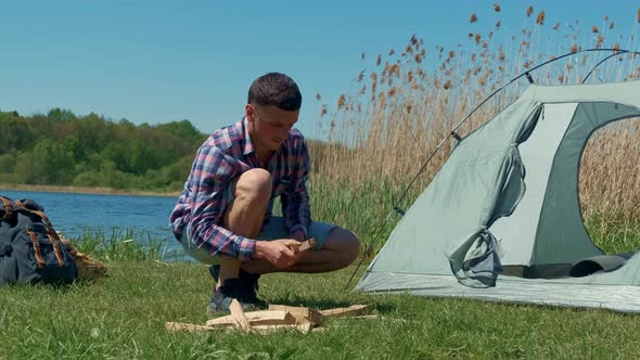 Young Handsome Man Chopping Wood with an Ax in Nature for Making a Fire in a Camping alt
