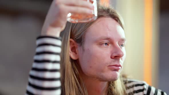 Closeup of Drunk Hipster with Drink Glass Sitting at Bar Counter Sighing and Looking Around alt