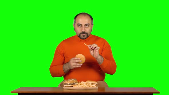 Man at a Table with a Plate of Fast Food Enjoys Eating a Burger and French Fries, Green Screen alt