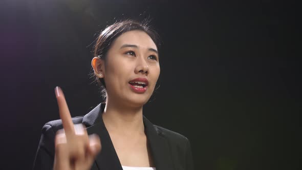 Side View Of Asian Speaker Woman In Business Suit Showing Index Fingers Up While Speaking On Stage alt